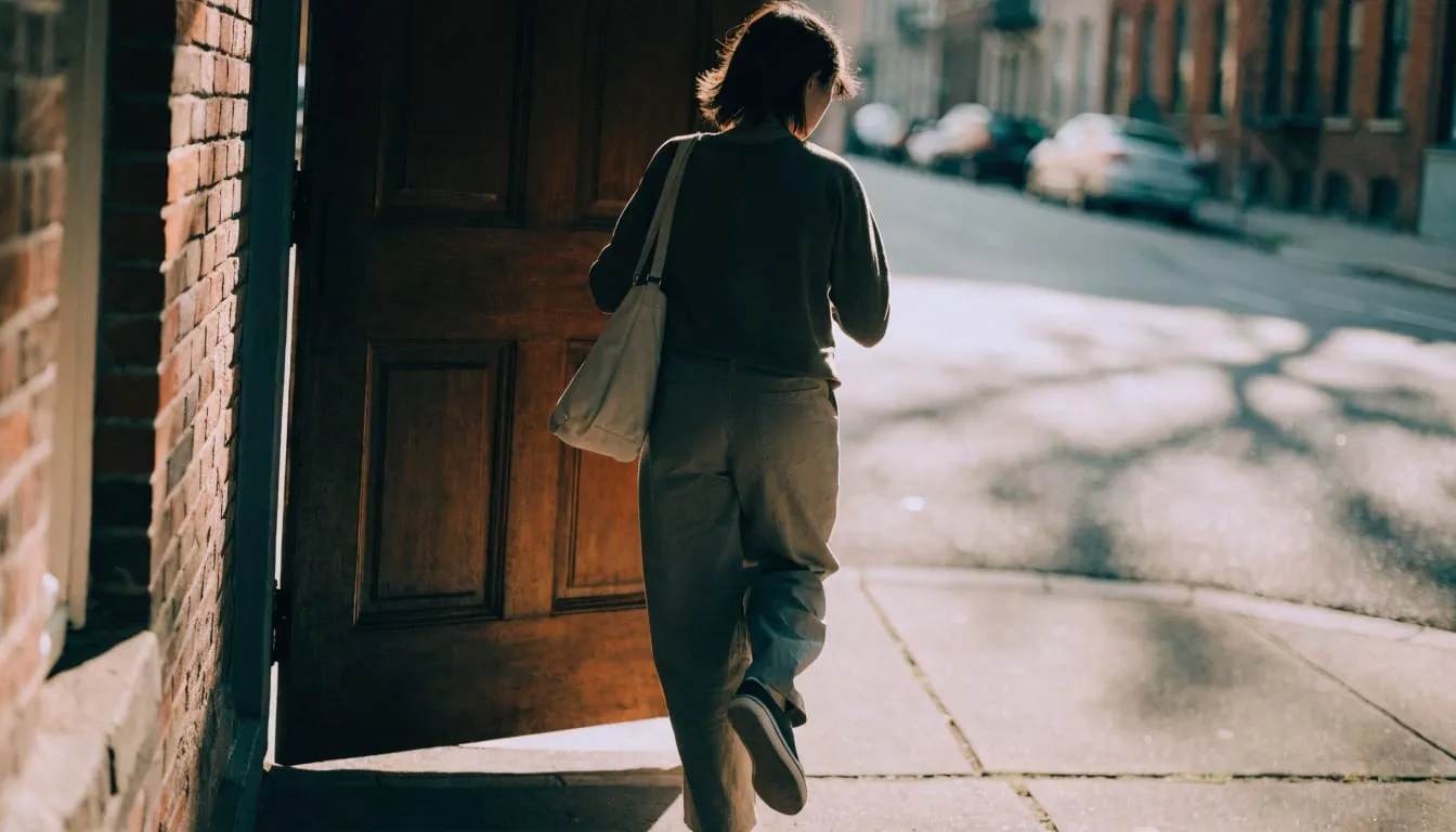 A person steps out of a rowhouse front door into bright morning light