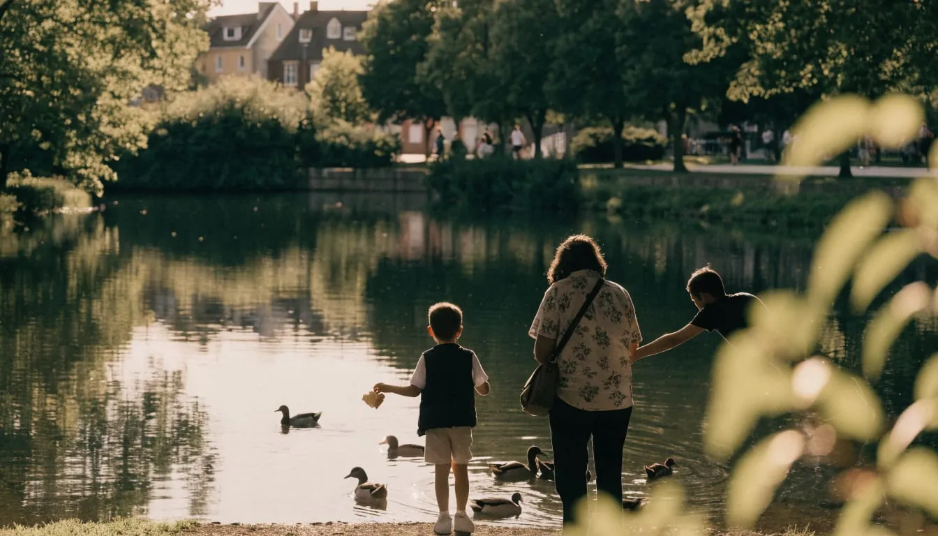 A family feeds ducks beside a calm tree-ringed pond on a warm afternoon