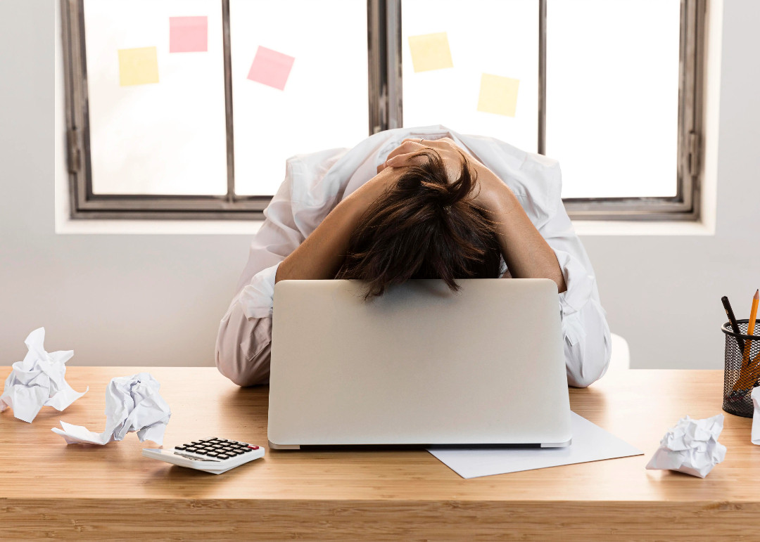 Someone rests their head on a desk behind a laptop, surrounded by crumpled paper and sticky notes.