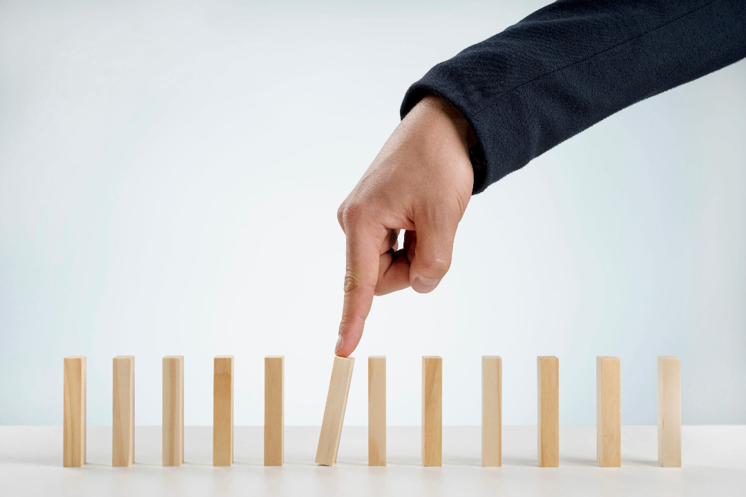 A hand stops one wooden block from toppling the rest of the row.