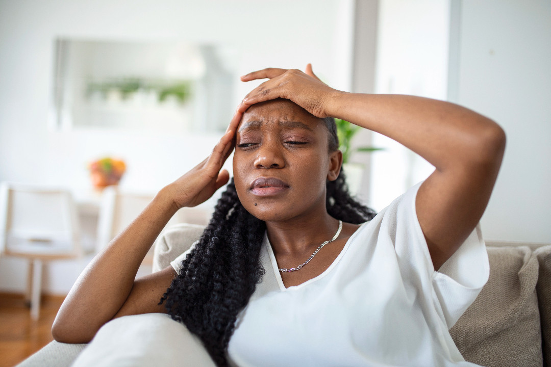 Someone sits on a sofa with eyes closed and both hands pressed to their head, looking overwhelmed.