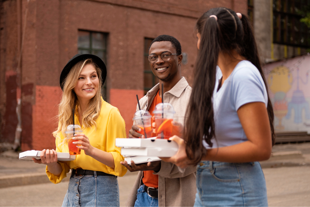 Three friends stand outside talking, holding cold drinks and pizza boxes.