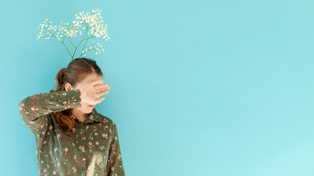 Someone covers their eyes with one hand, small white flowers rising behind their head.