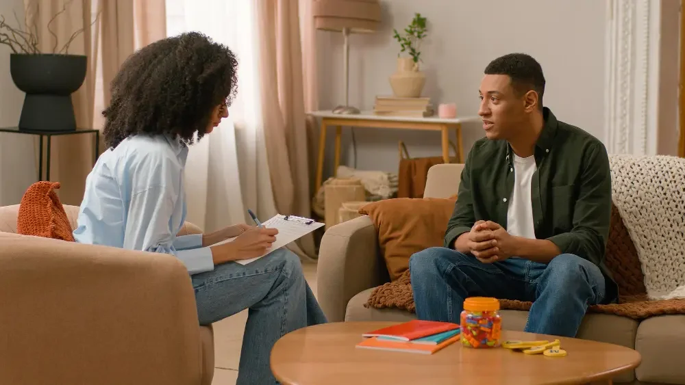 A clinician with a clipboard listens as a client talks during a therapy session in a warm living room.