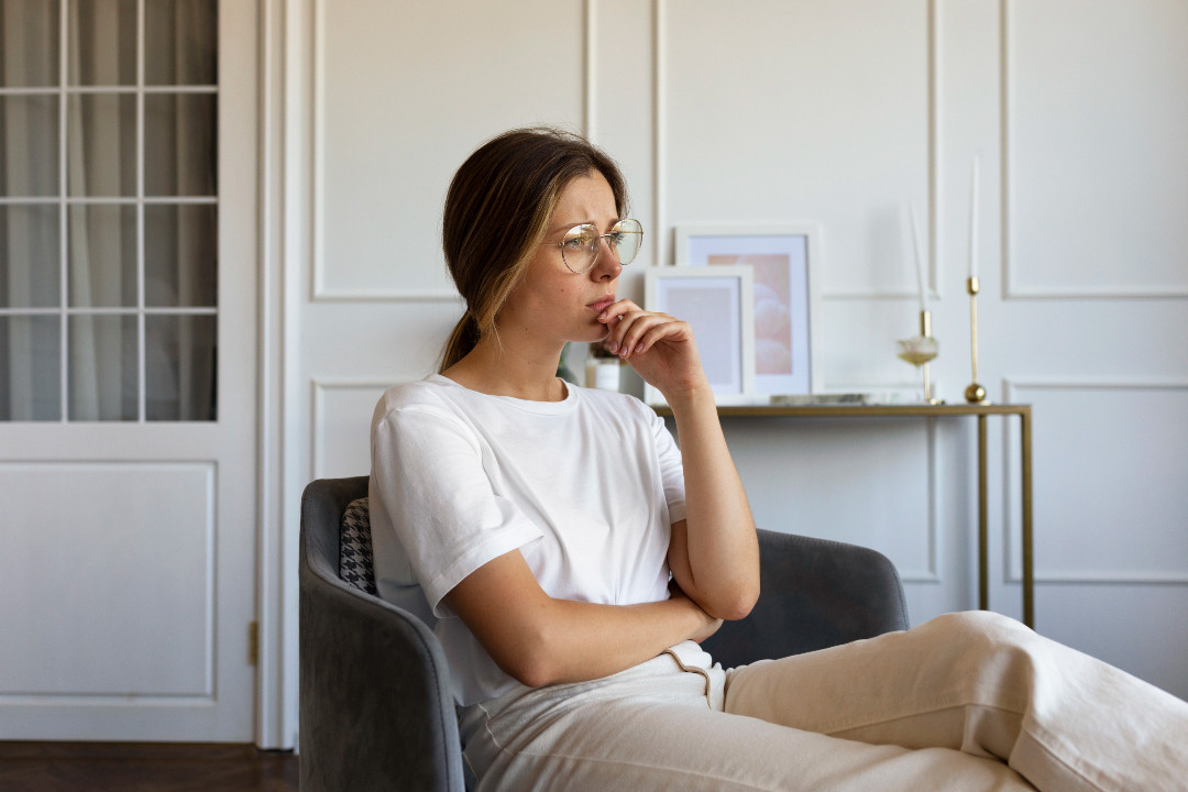 Someone sits alone in an armchair, hand to chin, looking away in quiet distress.