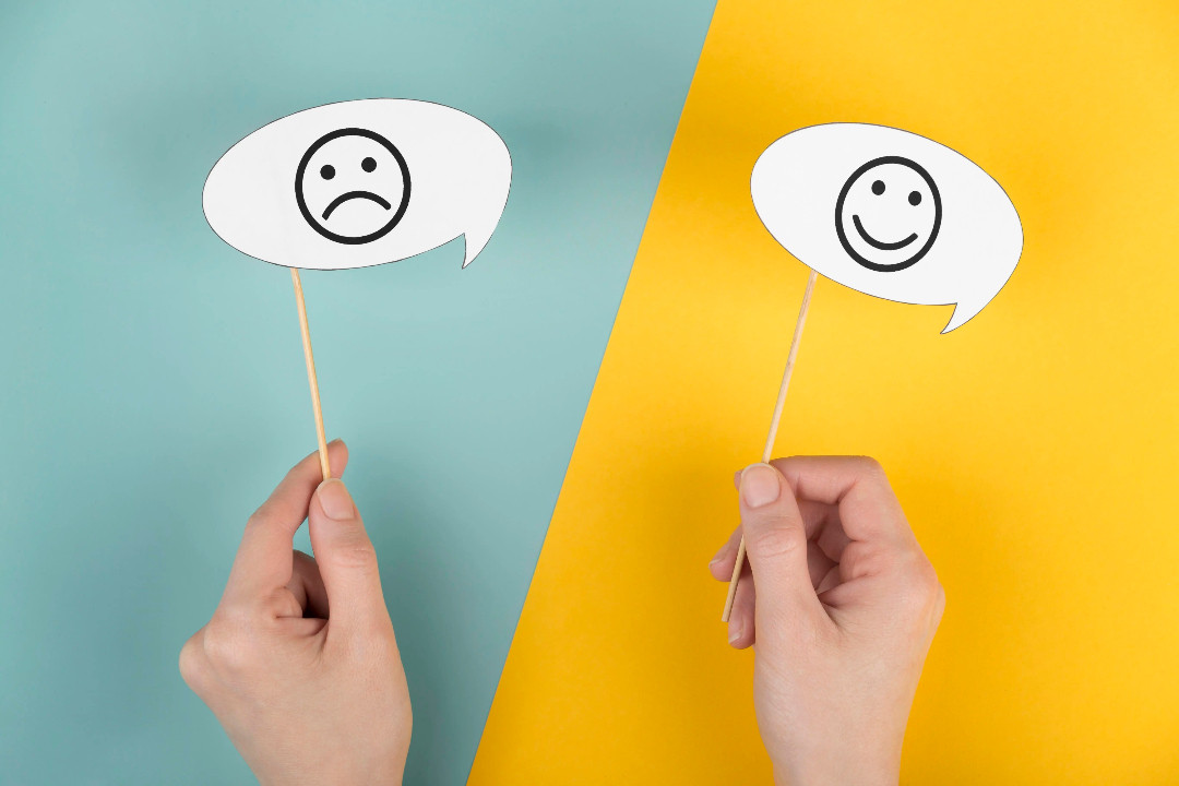 Two hands hold paper speech bubbles — a frowning face on teal, a smiling face on yellow.